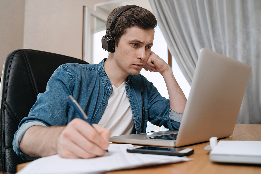 Serious young man in headphones looking at a laptop and writing on paper with a pen