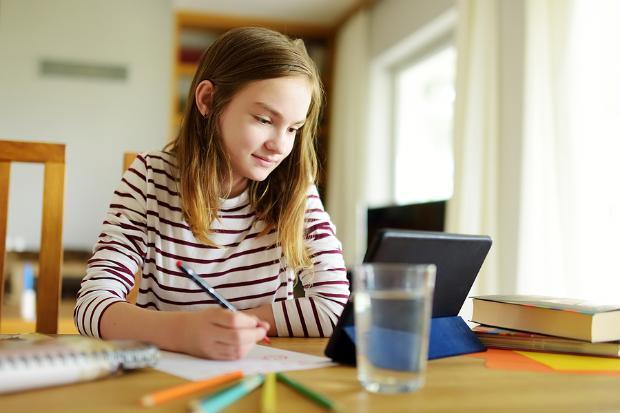 Girl in striped top writing at a desk, with a tablet, glass of water, and books nearby.