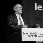Ian Wall speaking at a podium with a microphone, in front of a screen that says learning on screen