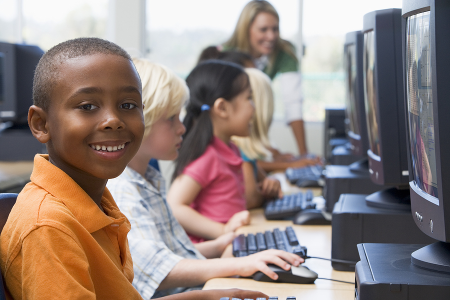 Smiling boy in orange polo shirt at a computer terminal, with other children and a teacher in a classroom.