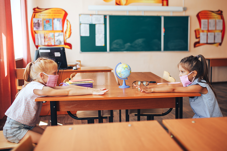 Two young schoolgirls in pink face masks sit at separate desks, facing each other with hands outstretched.