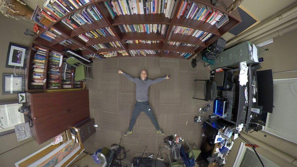 A man lying on his back on a patterned carpet, arms and legs outstretched, surrounded by bookshelves.