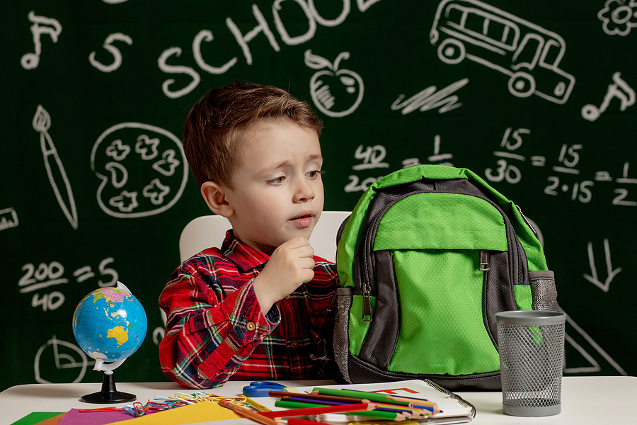 Young boy in a plaid shirt looking into a green backpack, with a chalkboard and school supplies
