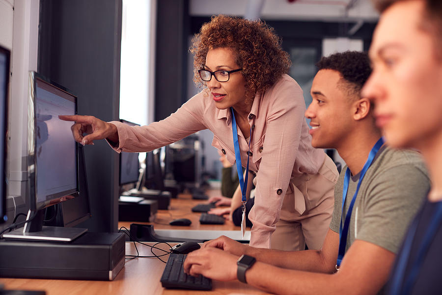 Teacher points at a computer screen while a student types on a keyboard in a classroom.