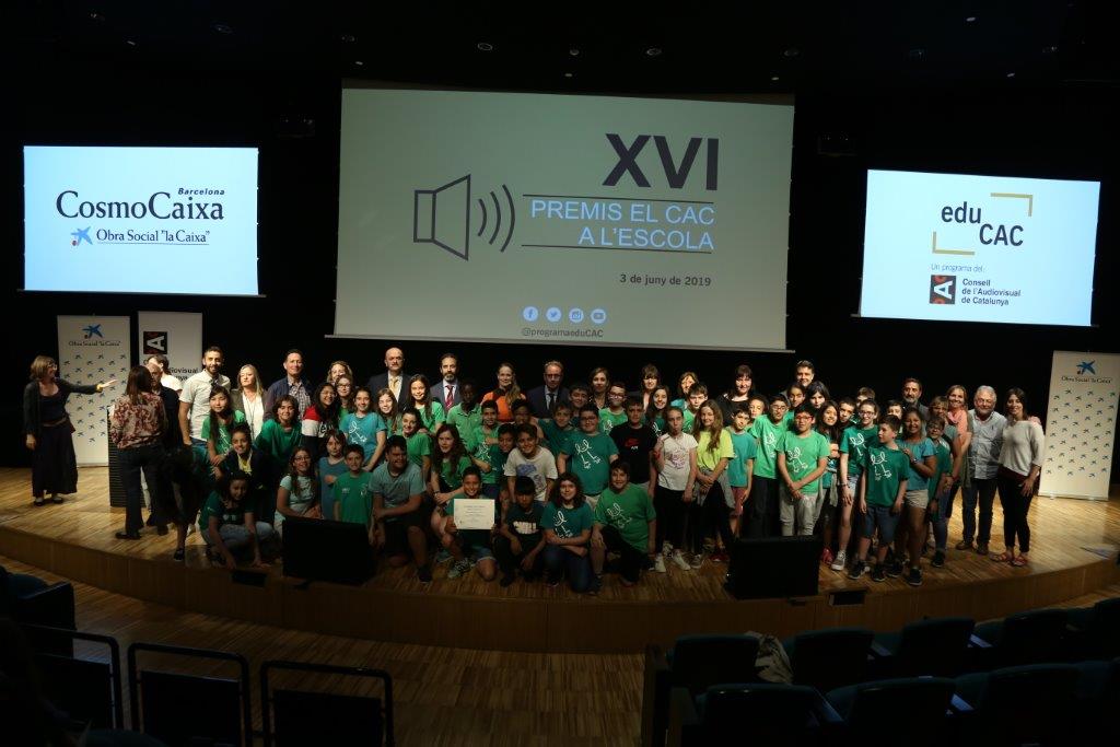 Group photo of children and adults on a stage, with screens showing event logos and titles in the background