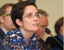 A woman with short dark hair and glasses speaks into a microphone at a conference table