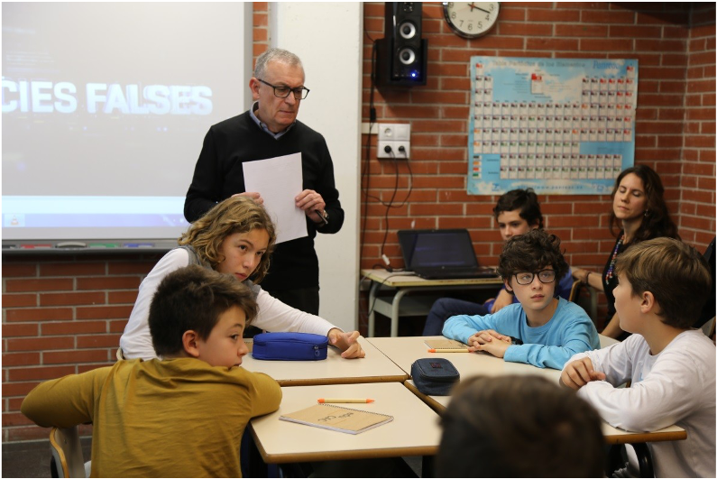 A man holding papers speaks to a group of students in a classroom with a periodic table on the wall.