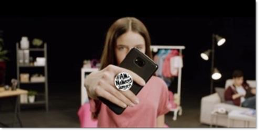 A young woman holds a smartphone with a pop socket that reads "#AM. Naked. Danger." towards the viewer.