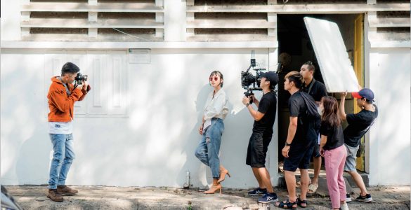 Image of a group of young people making a film, with one person taking a photo of the film crew, one holding the camera and another holding up a light reflector.