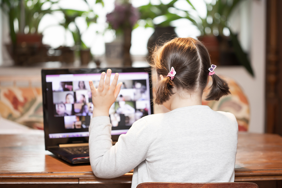 A young girl with pigtails raises her hand while looking at a laptop screen showing a video call.