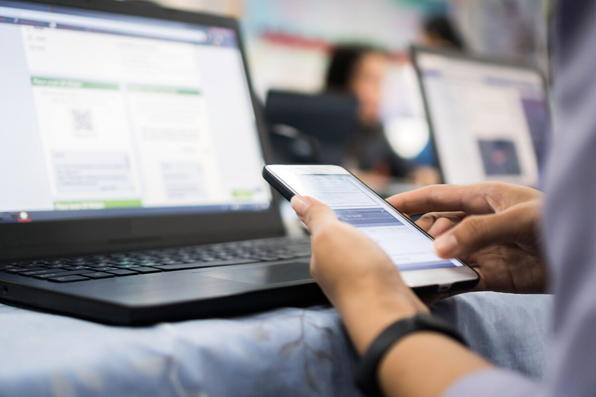 Person holding a smartphone, with a laptop displaying a website in the background.