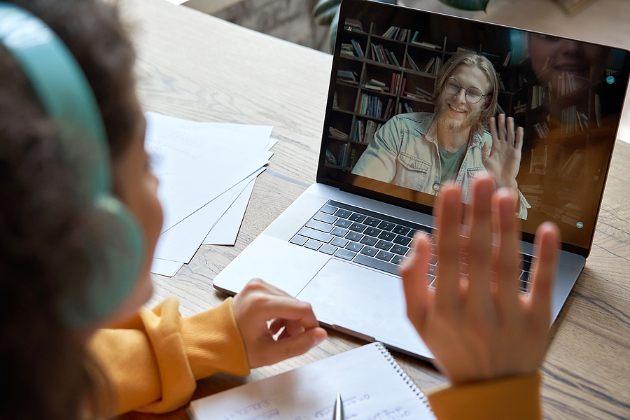 Student with headphones on, waving at a teacher on a laptop screen during an online class