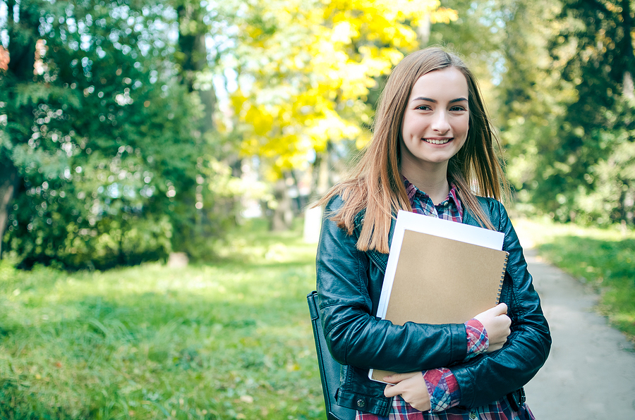Smiling female university student with long brown hair, wearing a leather jacket and plaid shirt, holding books