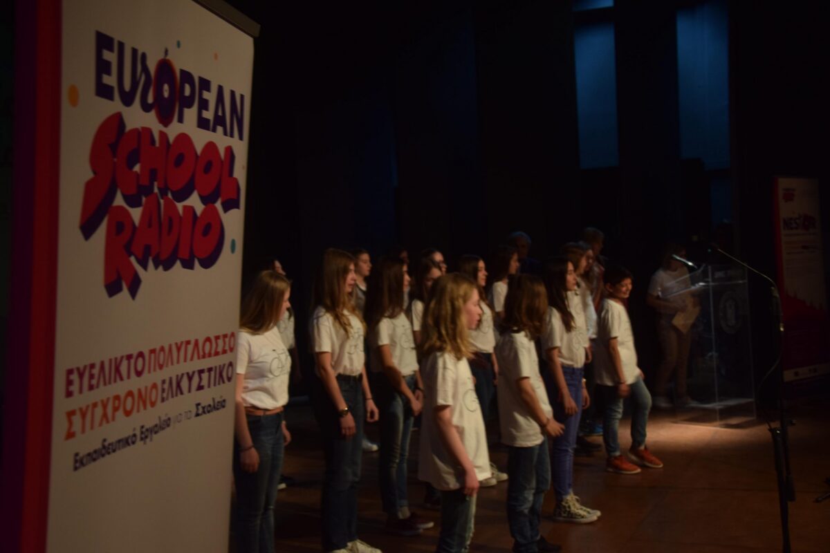 Students in white t-shirts and jeans standing on a stage, with a banner for European School Radio