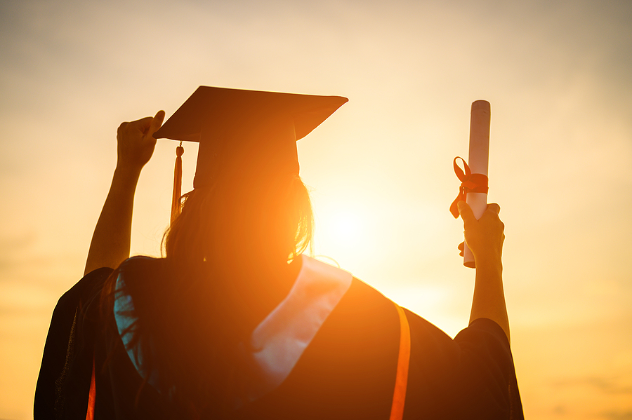 Graduate in cap and gown, seen from behind, holding a diploma aloft against a sunset sky.