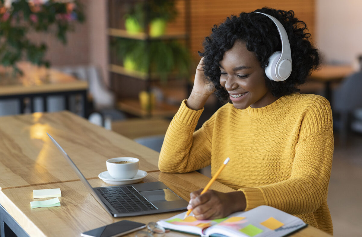 Smiling woman with headphones writing in a notebook, with a laptop and coffee on a wooden table.