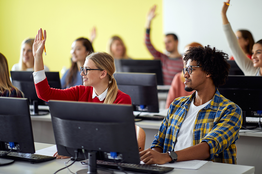 Students in a computer lab, some raising their hands, including a woman with glasses and a man with curly hair.