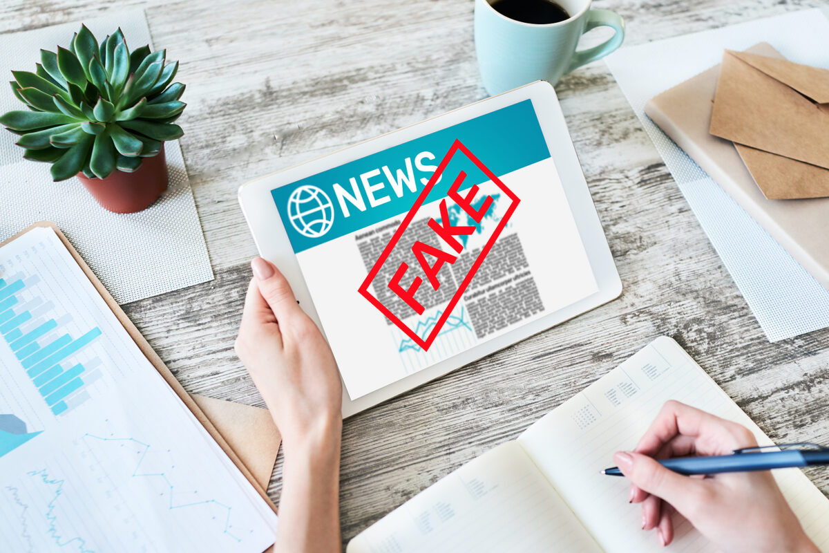 A person holding a tablet displaying a news article with a red 'FAKE' stamp over it, writing in a notebook.