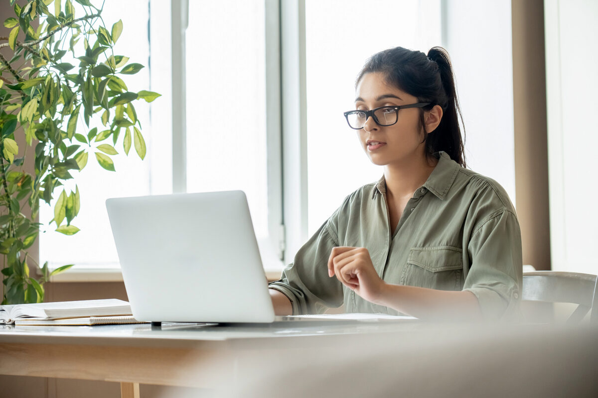 Young woman with glasses and a ponytail, wearing a khaki shirt, looking at a laptop and speaking.