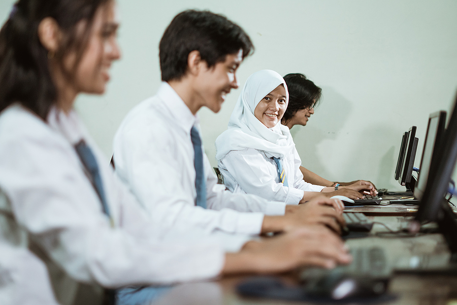 Four high school students in uniform using computers in a classroom, one girl in a hijab smiles at the camera.