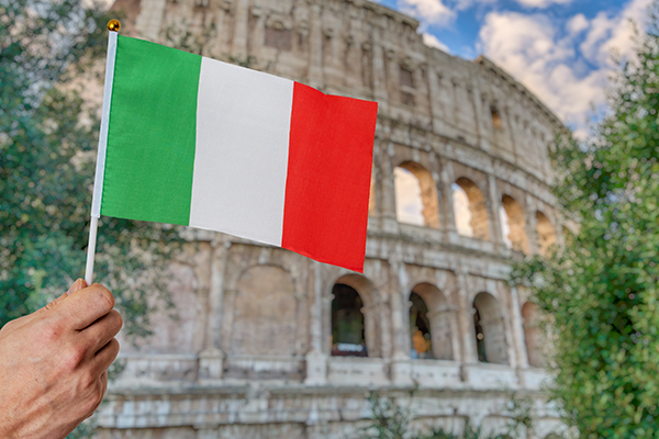 A hand holding a small Italian flag with the Colosseum in Rome visible in the blurred background.