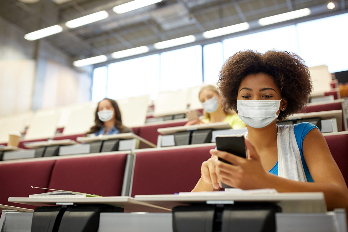 Students in a lecture hall wearing face masks and social distancing, one student uses a phone.