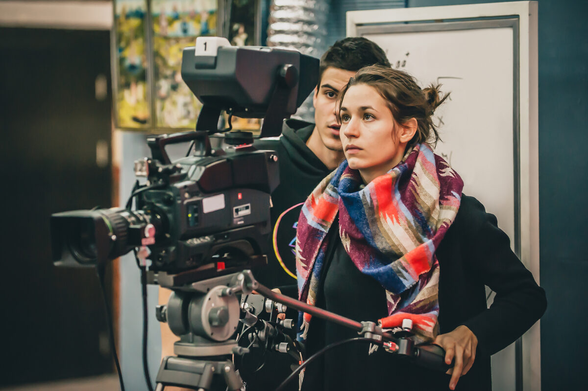 Two young people, one operating a professional video camera on a tripod, the other looking intently.