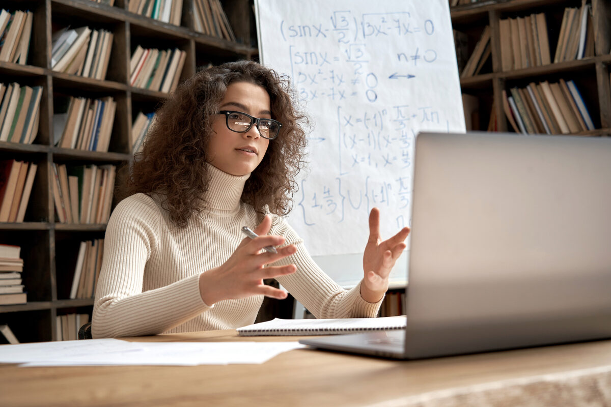 Young woman with curly hair and glasses talking on a video call, with a whiteboard of maths equations behind her.