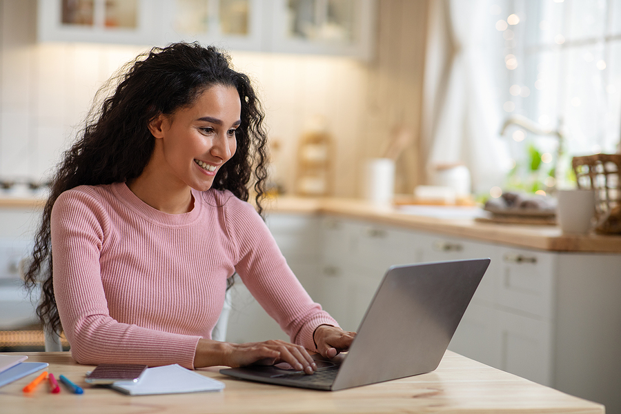 A smiling woman with long dark curly hair types on a laptop at a wooden table in a kitchen.