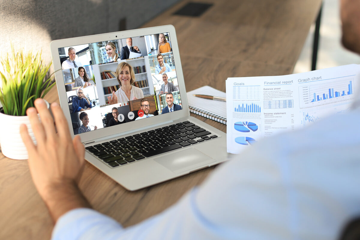 A person's hand waves at a laptop screen showing a video conference with 12 participants.
