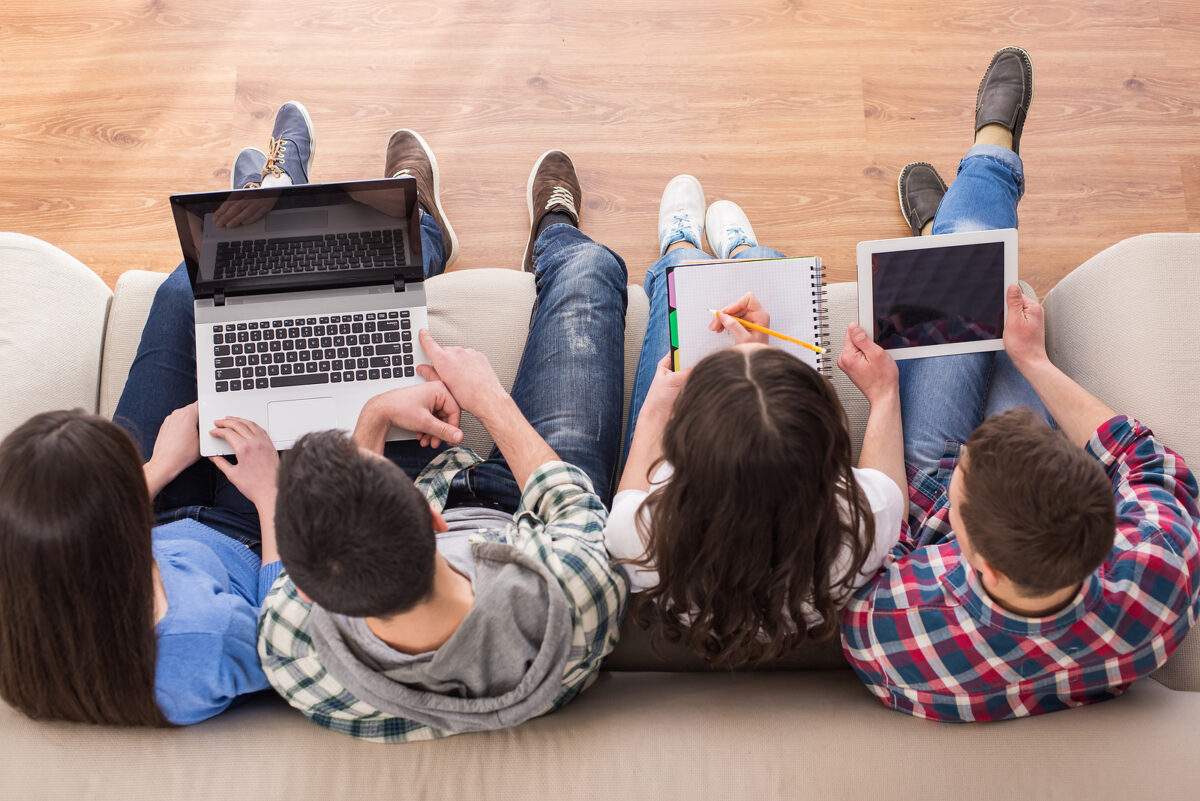 Four students sitting on a sofa, using a laptop, writing in a notebook, and using a tablet.