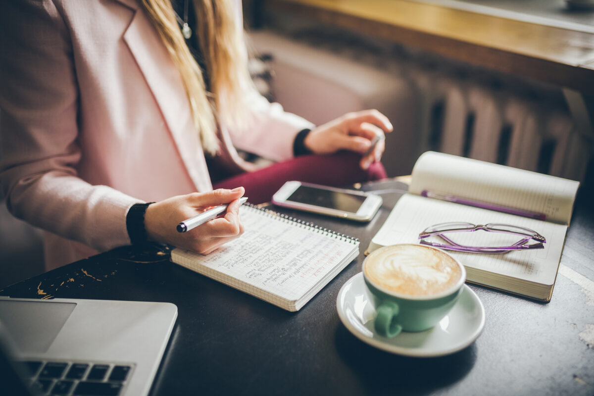 Woman in a pink blazer writing in a notebook, with a laptop, phone, coffee, and open book on a table.