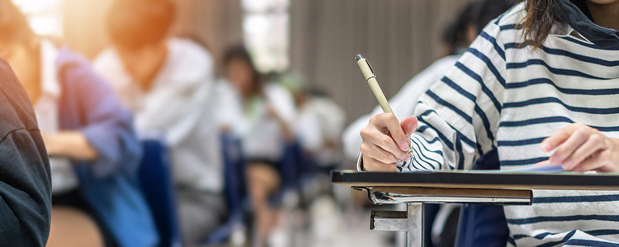 A student in a striped top writing with a pen at a desk in a classroom, other students blurred in background.