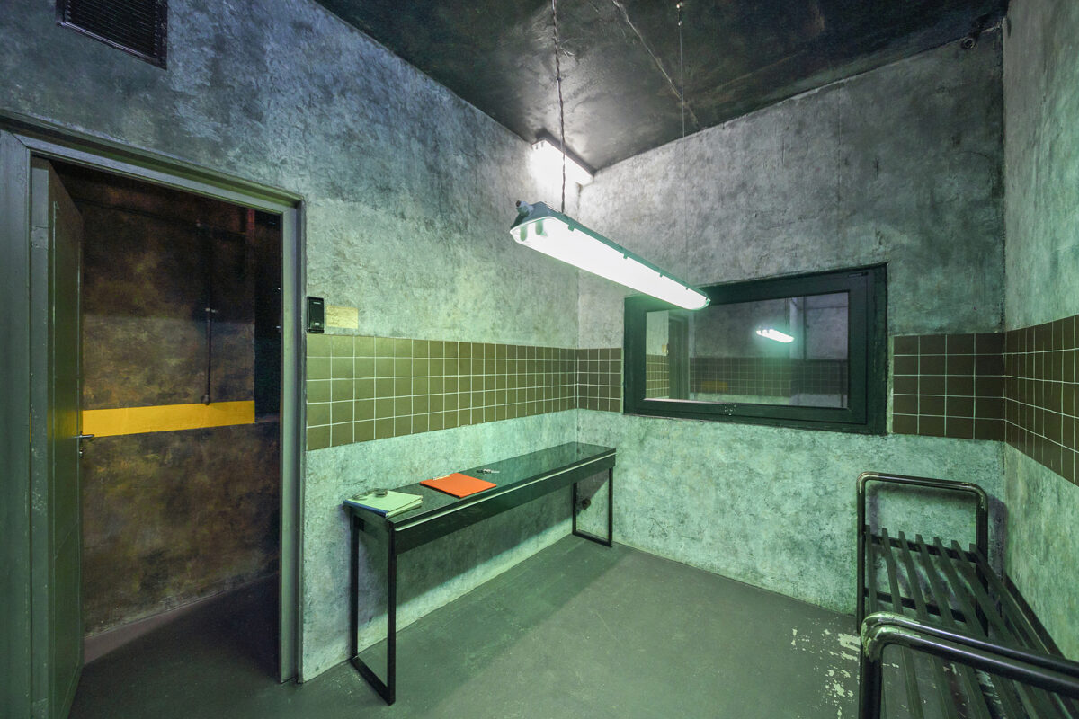 A dimly lit, grey concrete room with a long fluorescent light, a window, a desk, and a metal bed frame.