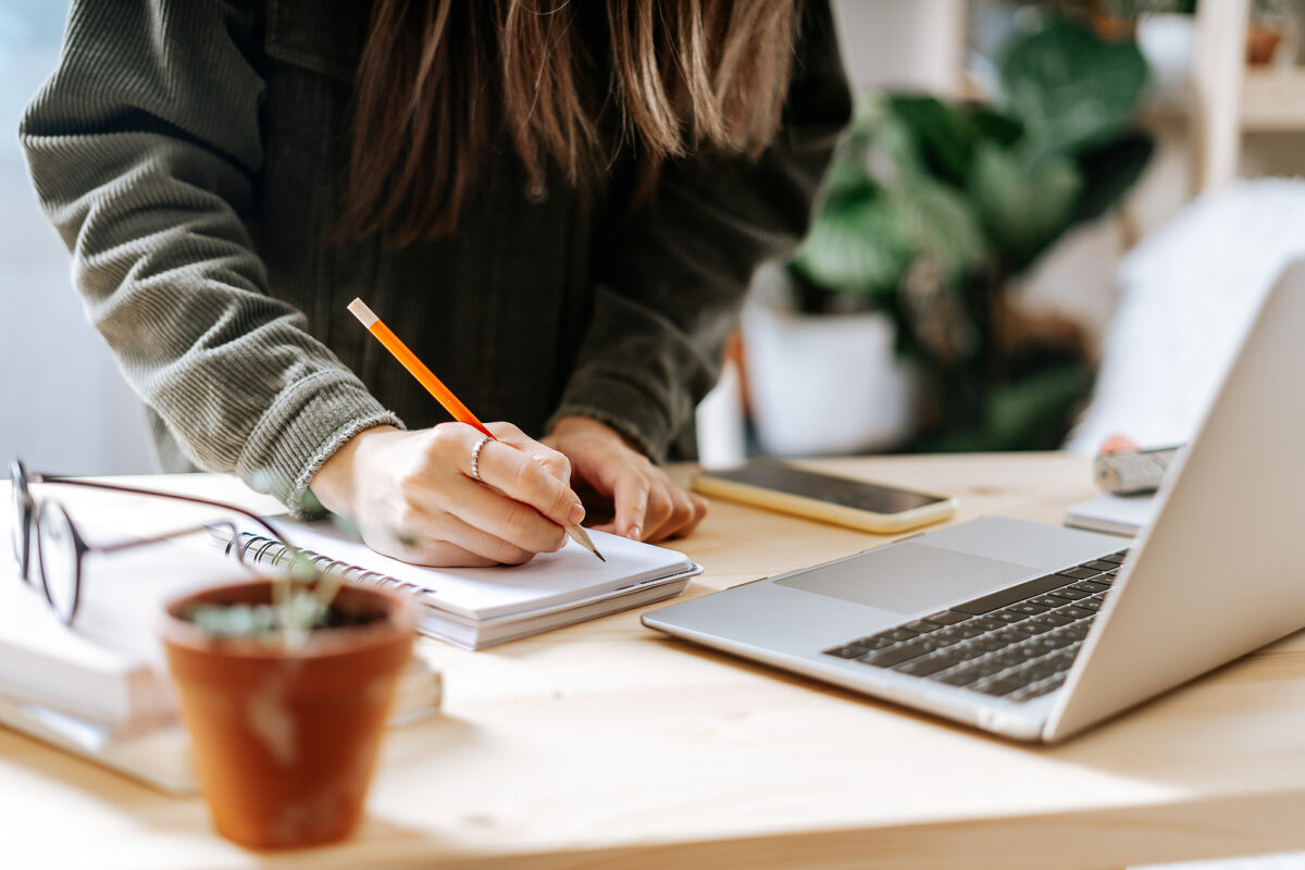 Person in a green corduroy shirt writing in a notebook with a pencil, next to a laptop and phone