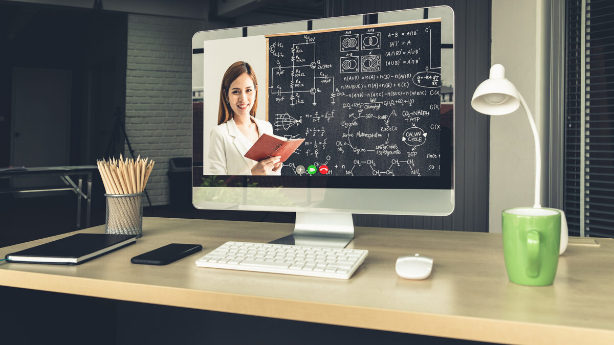 A woman teaching online, visible on a computer screen, with a blackboard full of equations behind her.
