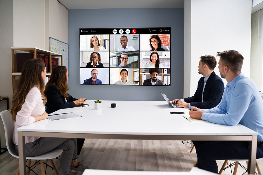 People in a meeting room looking at a large screen showing a video conference with nine participants.
