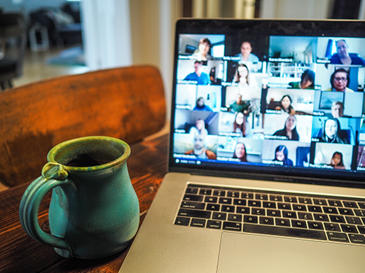 A laptop screen shows a video conference with many participants, next to a teal mug on a wooden table.