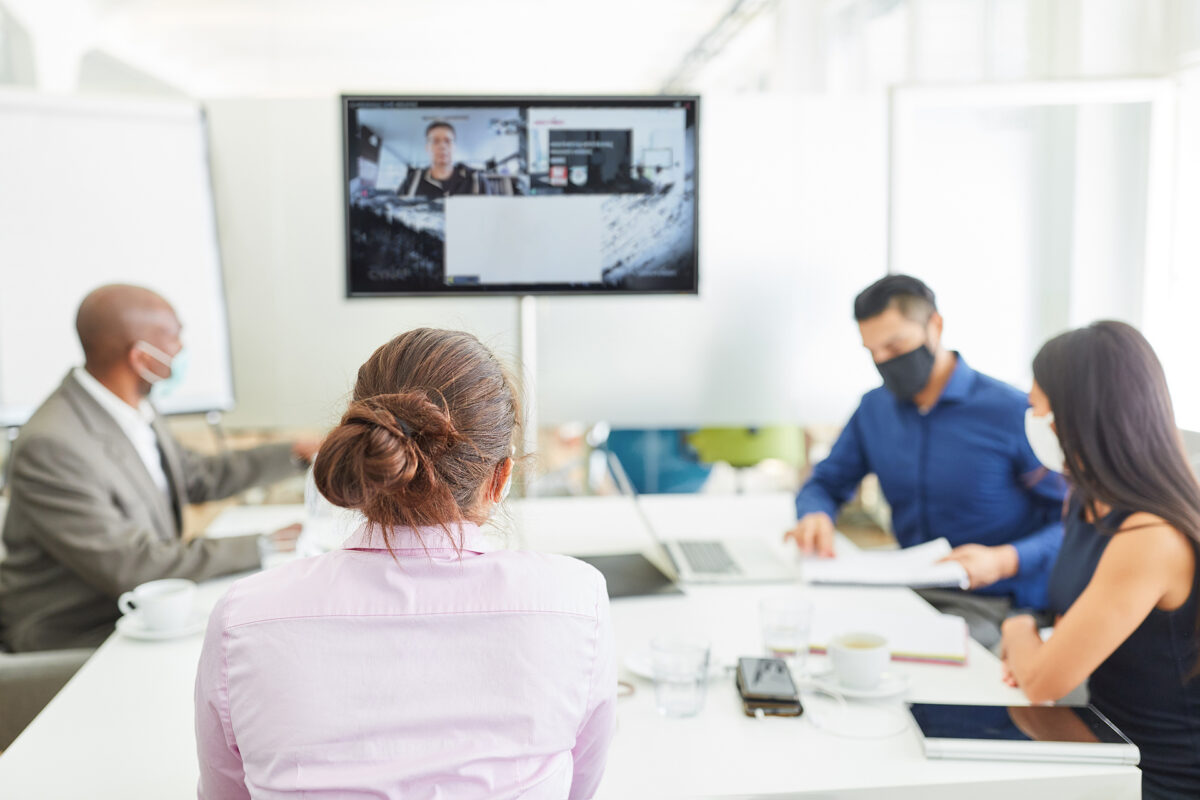People in a hybrid meeting, some in the room wearing masks, others on a screen via video call.