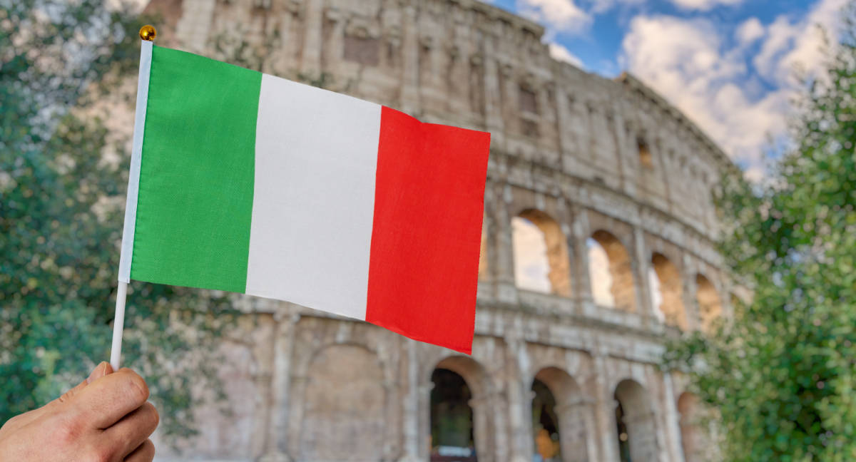 A hand holding a small Italian flag with the Colosseum in Rome visible in the blurred background.