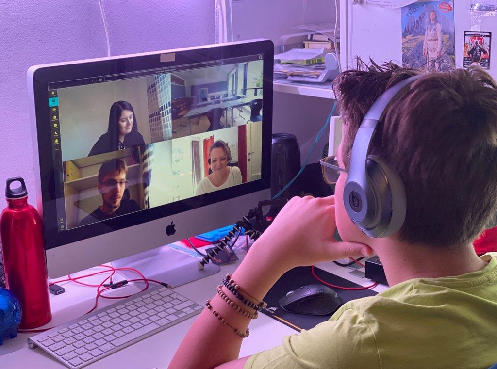 A student wearing headphones watches a video conference with four other people on a desktop computer.