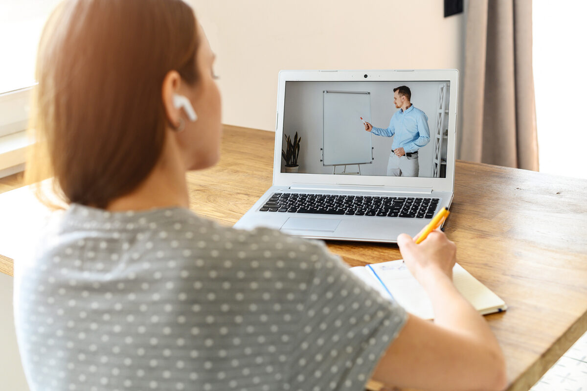 A woman with earbuds watches a man on a laptop screen, taking notes with a yellow pen.