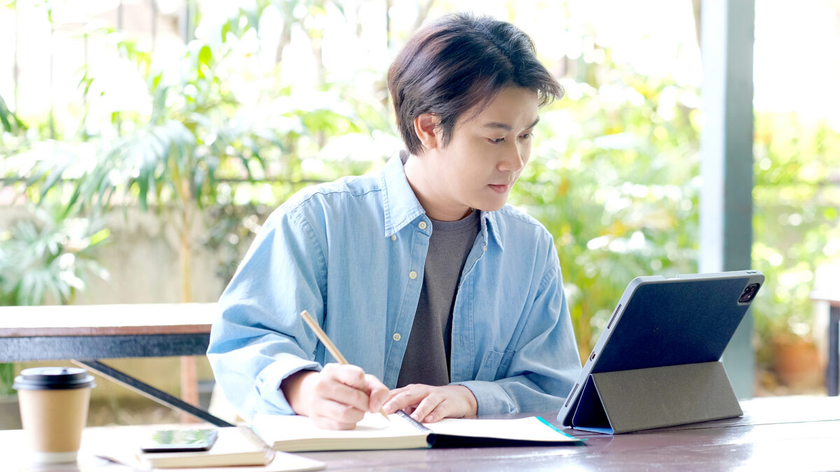 Young person in a blue shirt writing in a notebook while looking at a tablet, with a coffee cup nearby.