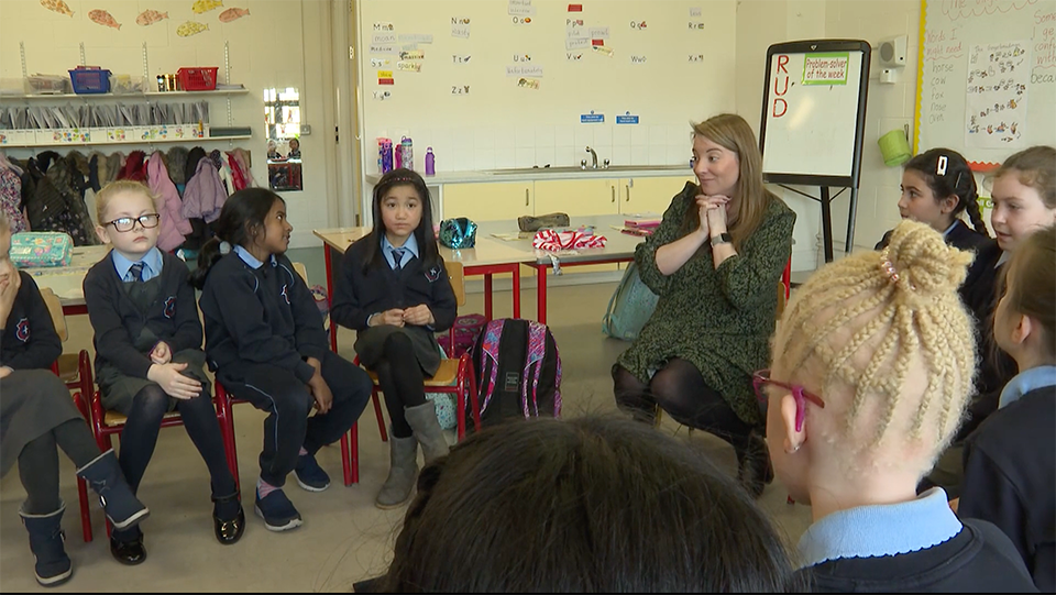 A teacher sits on the floor with her hands clasped, surrounded by primary school children in uniform.