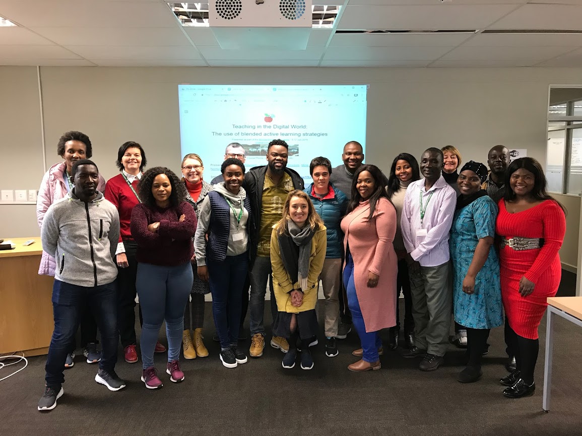 A group of 20 people, men and women, standing and smiling in a classroom with a projector screen.
