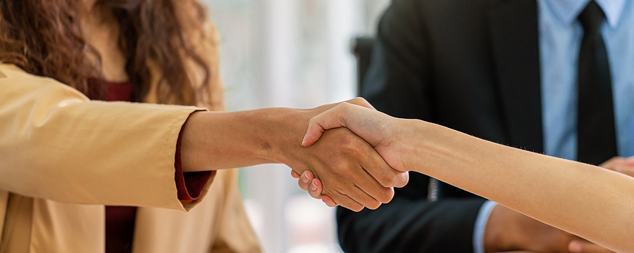 Two people shaking hands, one in a beige jacket and the other in a suit and blue shirt.