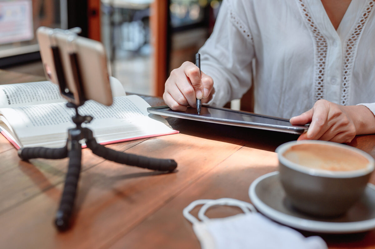 Person using a stylus on a tablet, with a book, smartphone on tripod, coffee, and face mask nearby.