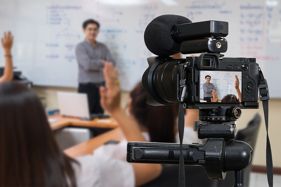 A camera on a tripod records a male teacher at a whiteboard and students raising hands in a classroom.
