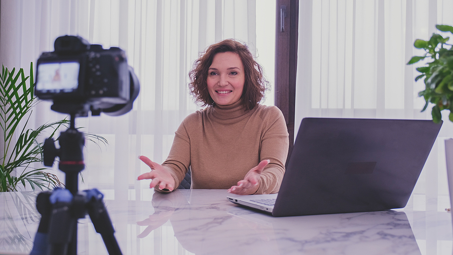 A smiling woman in a brown turtleneck records a video at a desk with a camera and laptop.