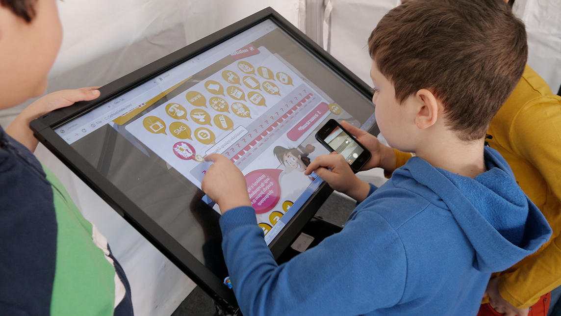 Two boys interacting with a large touchscreen displaying a media literacy game, one holding a smartphone.